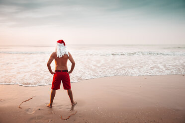 Thailand, Rückenansicht eines als Weihnachtsmann verkleideten Mannes, der bei Sonnenuntergang am Strand steht und auf den Horizont schaut - HMEF00213