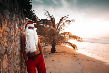 Thailand, man dressed up as Santa Claus leaning against wall on the beach at sunset - HMEF00214