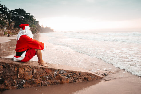 Thailand, als Weihnachtsmann verkleideter Mann sitzt bei Sonnenuntergang auf einer Mauer am Strand - HMEF00216