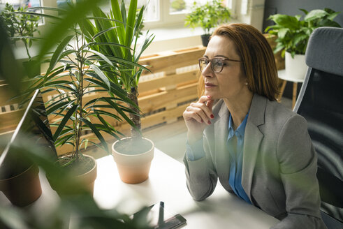 Reife Geschäftsfrau, die in einem nachhaltigen Büro arbeitet, mit Pflanzen auf ihrem Schreibtisch - MOEF02025