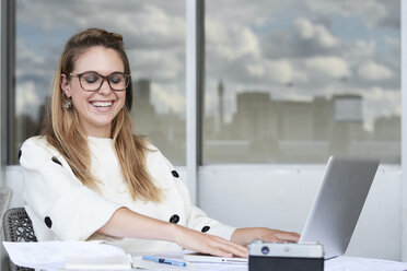 Portrait of laughing young businesswoman working on laptop in office - IGGF00799