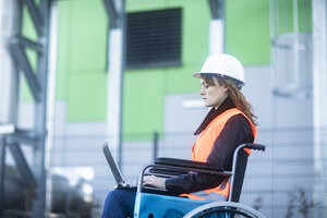 Junger Techniker mit Schutzhelm und -weste im Rollstuhl bei der Arbeit am Laptop im Freien - SGF02247
