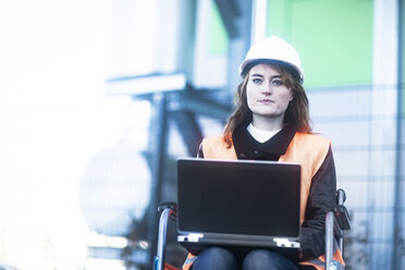 Portrait of young technician with safety helmet and vest in wheelchair working on laptop outdoors - SGF02248