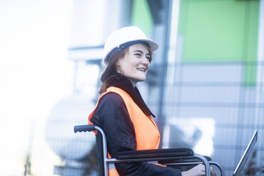 Young technician with safety helmet and vest in wheelchair working on laptop outdoors - SGF02250