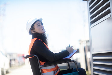 Young technician with safety helmet and vest in wheelchair working outdoors - SGF02253