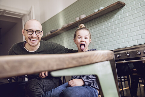 Vater und Tochter sitzen in der Küche, das Mädchen zieht lustige Grimassen, lizenzfreies Stockfoto