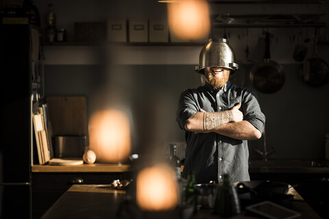 Mann steht in der Küche und trägt ein Sieb als Helm, lizenzfreies Stockfoto