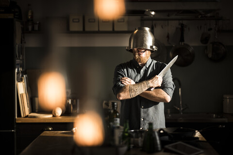 Mann mit Küchenmesser in der Küche stehend, mit Sieb als Helm, lizenzfreies Stockfoto