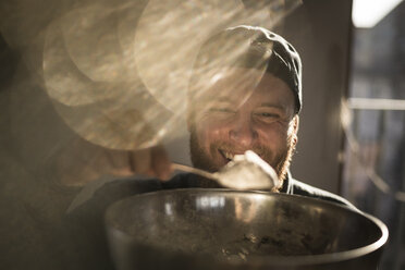Laughing man holding bowl in kitchen, adding ingredients to his dough - MJRF00051