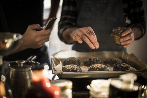 Young man taking pictures of friend, sprinkling sesame on burger buns on a baking tray stock photo