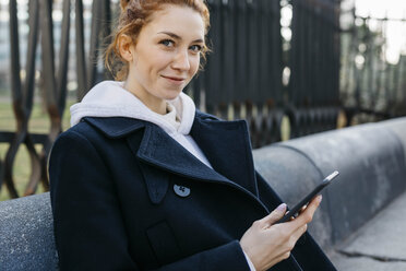 Porträt einer lächelnden jungen Frau, die auf einer Bank sitzt und ein Mobiltelefon hält - JRFF02674