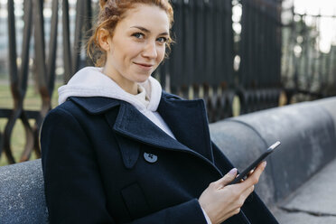 Portrait of smiling young woman sitting on a bench holding cell phone - JRFF02674