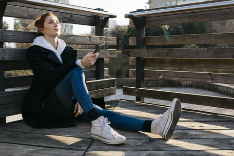 Junge Frau, die sich im Freien ausruht und ein Mobiltelefon hält, lizenzfreies Stockfoto