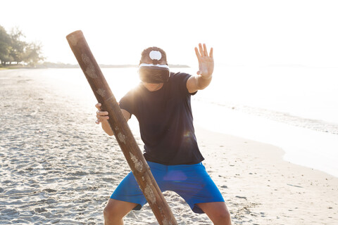 Mann mit Holzstab und VR-Brille am Strand, lizenzfreies Stockfoto