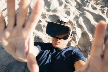 Man wearing VR glasses lying in sand on the beach - HMEF00226