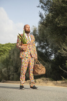 Man wearing suit with colourful polka-dots standing on country road with travelling bag and potted plant - KBF00508