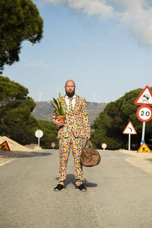 Man wearing suit with colourful polka-dots standing on country road with travelling bag and potted plant - KBF00509