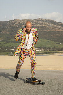 Smiling bald man on the phone wearing colourful suit while skateboarding on road - KBF00515