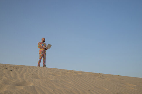 Mann mit Laptop und Anzug mit bunten Punkten steht auf einer Sanddüne und schaut in die Ferne - KBF00520