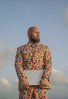 Portrait of bald man with beard wearing suit with colourful polka-dots holding laptop - KBF00523