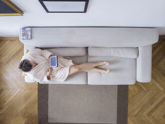 Mature woman in bathrobe sitting on the couch at home using digital tablet - PSIF00244