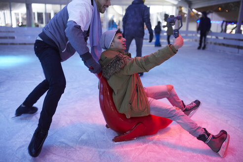 Glückliche verspielte Freunde mit einer Kamera beim Schlittschuhlaufen auf einer Eisbahn - ZEDF01919