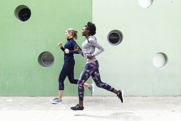 Zwei sportliche junge Frauen laufen zusammen in der Stadt an einer Mauer mit runden Fenstern vorbei - JSRF00137