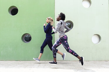 Zwei sportliche junge Frauen laufen zusammen in der Stadt an einer Mauer mit runden Fenstern vorbei - JSRF00138