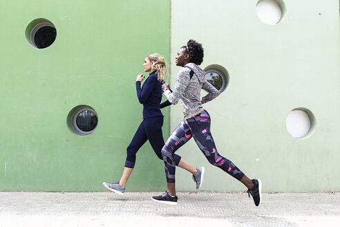 Two sporty young women running together in the city passing wall with round windows - JSRF00138
