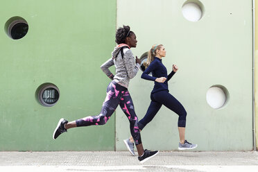 Two sporty young women running together in the city passing wall with round windows - JSRF00139