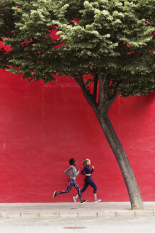 Two sporty young women running together in the city passing red wall - JSRF00140