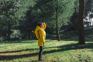 Mädchen mit gelbem Regenmantel und gelbem Rucksack steht auf einer Wiese im Sonnenlicht - ERRF00769