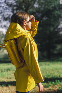 Mädchen mit gelbem Regenmantel und gelbem Rucksack steht im Sonnenlicht auf einer Waldlichtung - ERRF00771