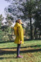 Girl wearing yellow raincoat and yellow backpack standing on a meadow enjoying sunlight - ERRF00772