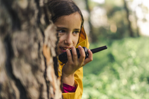 Portrait of girl with walkie-talkie hiding behind tree trunk - ERRF00780