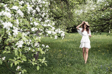 Young woman wearing white dress and floppy hat walking barefoot in garden with blossoming apple trees - WFF00017