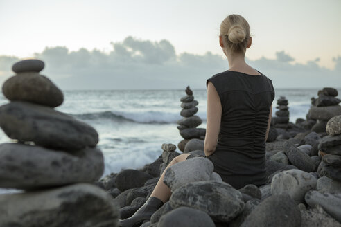 Spain, Tenerife, Costa Adeje, woman sitting between cairns at the coast - PSTF00304