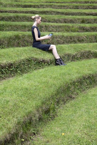 Italy, Alto Adige, Lana, woman sitting on grass-covered steps of natural open air theater using tablet stock photo