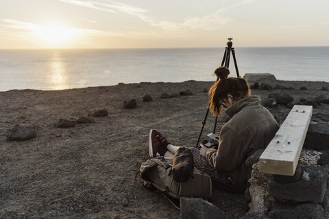 Spanien, Kanarische Inseln, Fuerteventura, junge Frau sitzt auf dem Boden und benutzt Smartphone und Digitalkamera, lizenzfreies Stockfoto