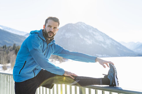 Deutschland, Bayern, sportlicher Mann streckt sich im Winter auf einem Brückengeländer - DIGF05978