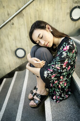 Porträt einer jungen Frau mit geschlossenen Augen, die auf einer Treppe sitzt und die Knie umarmt - DMOF00144