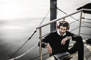 Businessman on a houseboat using laptop and smartwatch, wearing headphones - MJRF00077