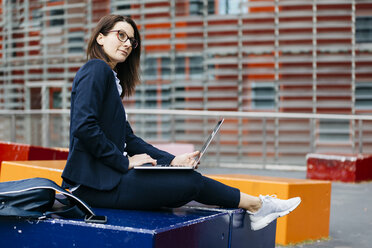 Businesswoman sitting outside office building in the city using laptop - JRFF02728