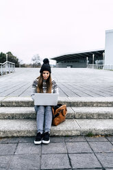 Junge Frau sitzt im Freien auf einer Treppe und benutzt einen Laptop - MGOF03956