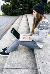 Junge Frau sitzt im Freien auf einer Treppe und benutzt einen Laptop - MGOF03960