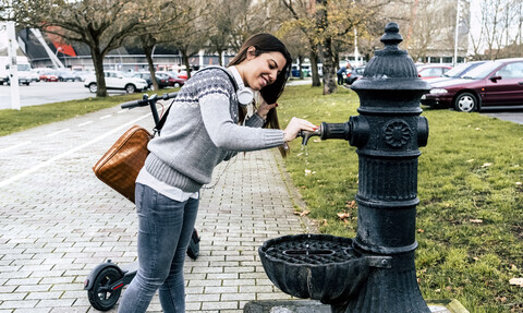 Lächelnde junge Frau mit Elektroroller trinkt Wasser aus einem Brunnen in der Stadt, lizenzfreies Stockfoto