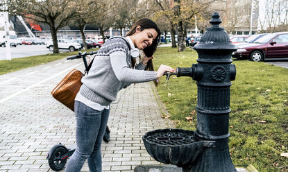 Lächelnde junge Frau mit Elektroroller trinkt Wasser aus einem Brunnen in der Stadt - MGOF03967