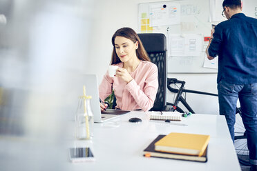 Woman working at desk in office with colleague in background - BSZF00994