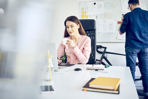 Frau arbeitet am Schreibtisch im Büro mit einem Kollegen im Hintergrund - BSZF00994