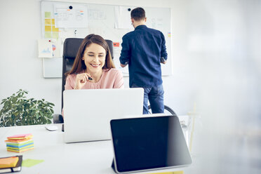 Woman working at desk in office with colleague in background - BSZF00995